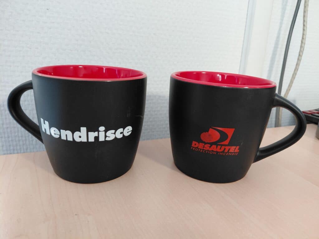 Deux tasses noires intérieur rouge marquées Hendrisce et Desautel Protection Incendie sur un bureau en bois.
